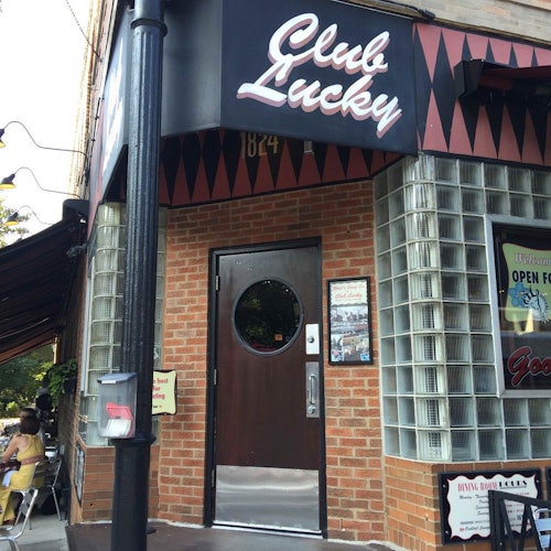 幸運俱樂部的入口，帶有圓形窗戶的門、紅磚外牆、遮陽篷和招牌。左側可見戶外座位區。
