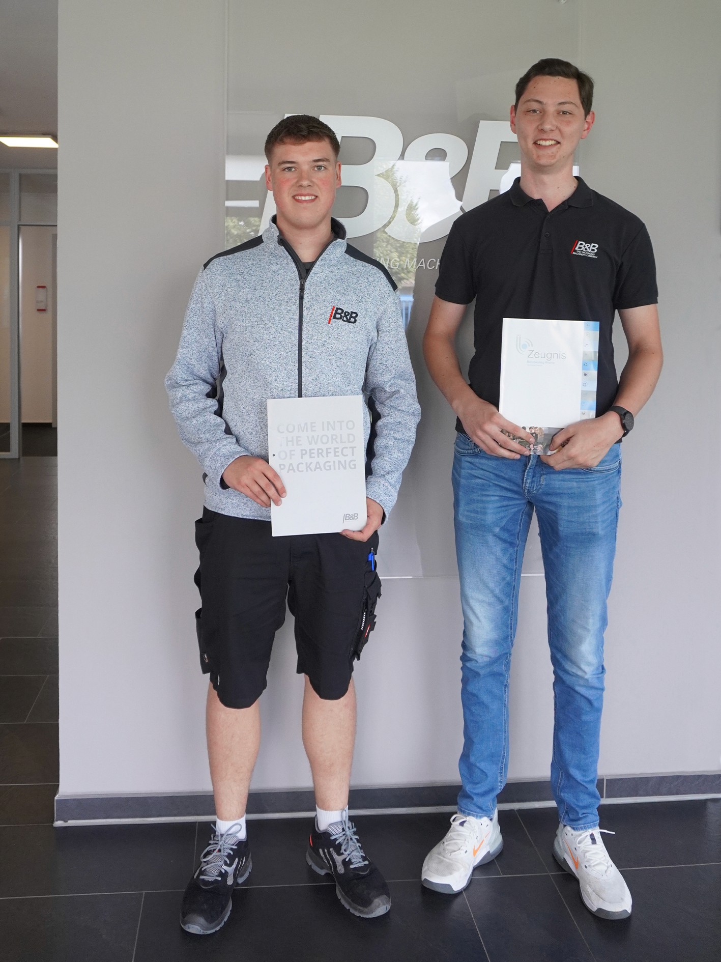 Zwei junge Männer mit Urkunden vor dem Firmenlogo. Erfolgreiche Ausbildung.