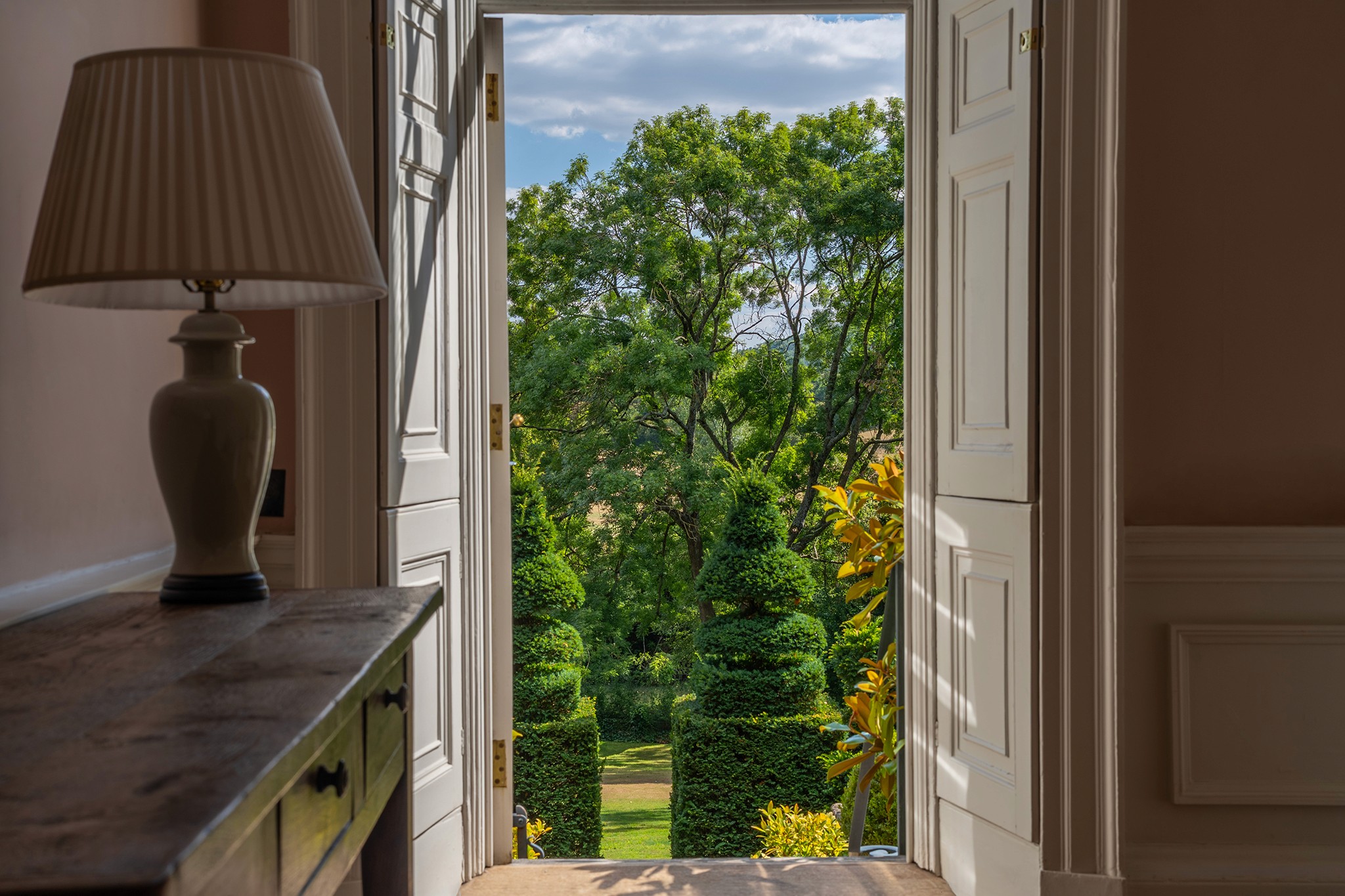 Antique console table and lamp framed by a shuttered doorway onto the topiary garden, Batheaston House, Bath