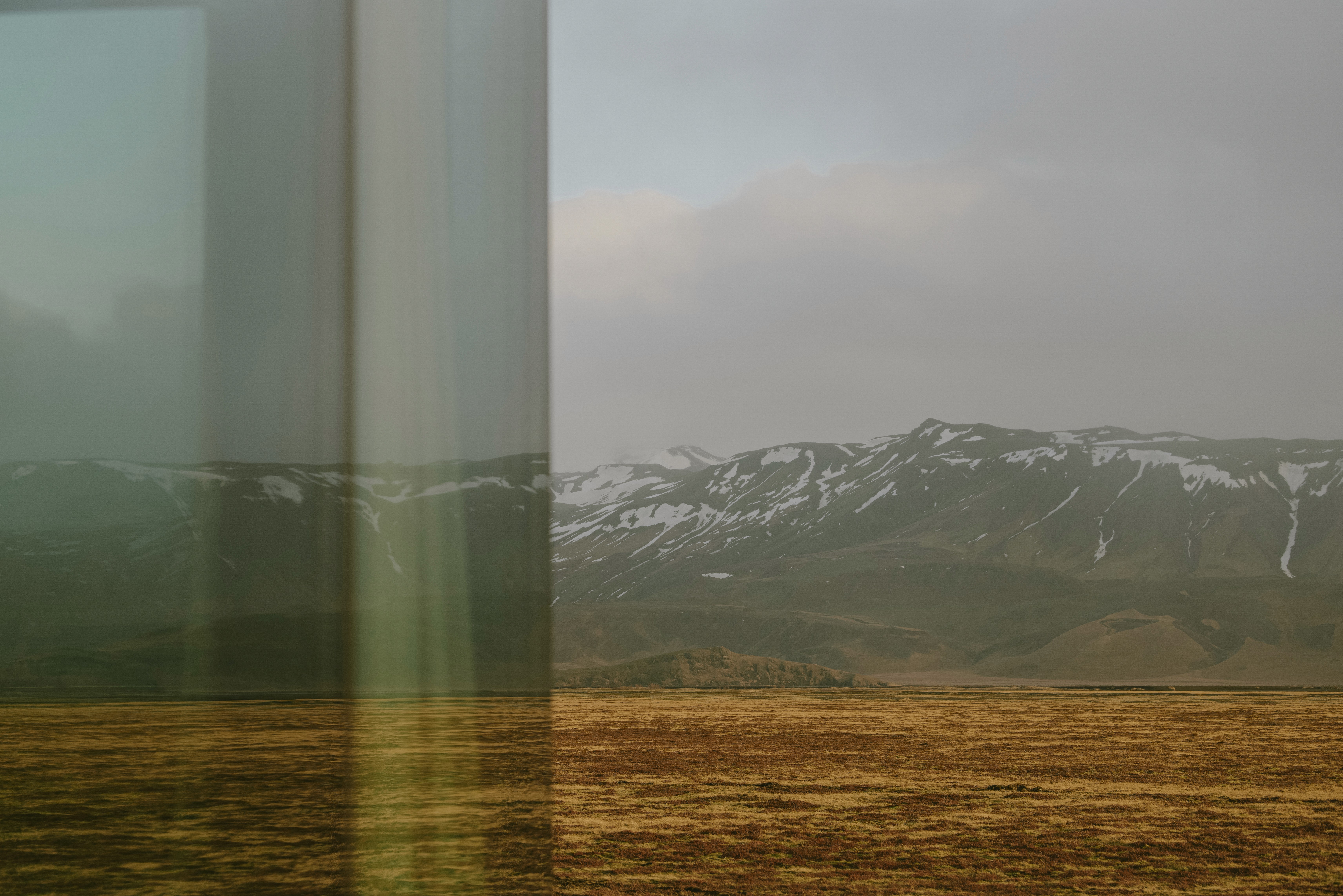 Panoramic view of a wide, open landscape with rolling plains and snow-capped mountains, seen through reflective floor-to-ceiling glass of a modern design-led cabin.