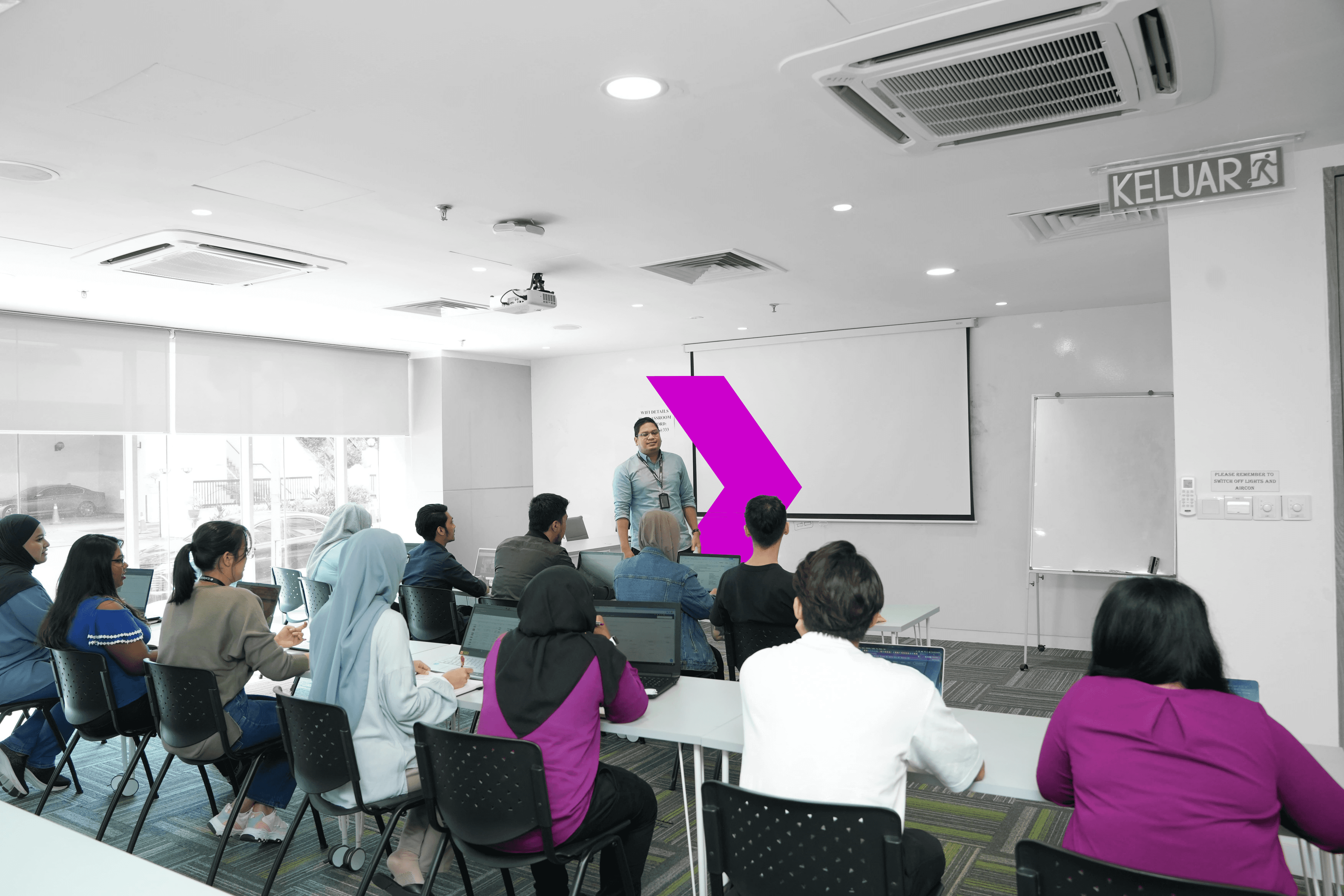 A group of students attentively listens to a speaker in a bright classroom with a projector and whiteboard.