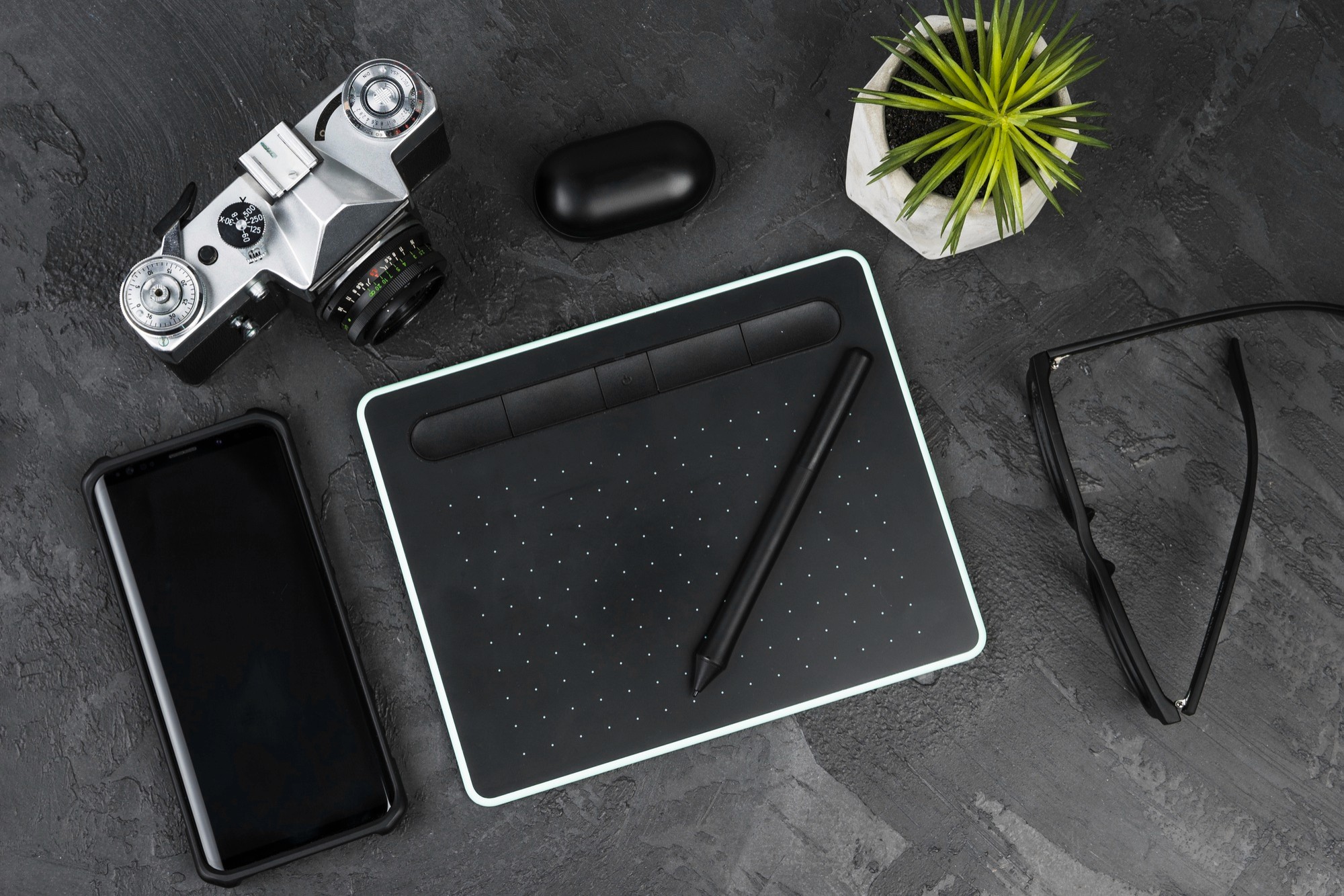 Overhead flat lay of a professional creative workspace featuring a black graphics drawing tablet with a stylus, a vintage-style camera, a smartphone, and a small succulent plant on a dark grey textured surface.