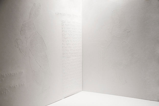 The image shows a white wall divided into two sections. On the left, there is a line drawing of a rabbit facing left. On the right, there is a line drawing of a figure resembling Alice from Alice in Wonderland, also facing left. Below the drawings, lines of text are embossed or printed on the wall. The floor beneath the wall is white.