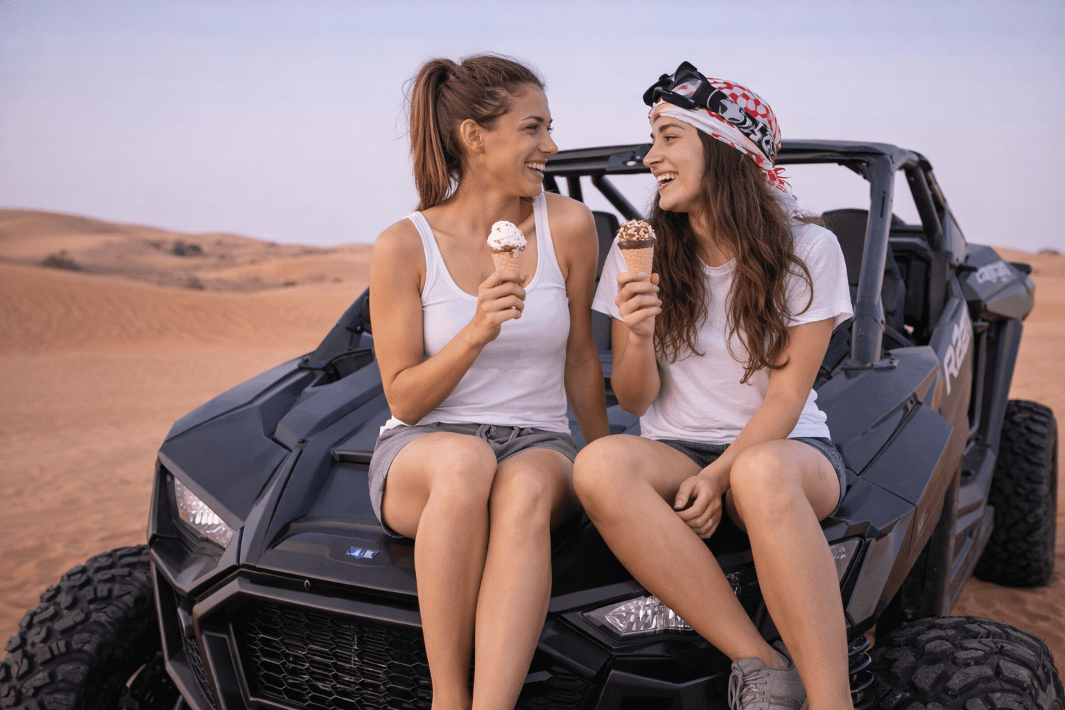 Two women eating ice cream while sitting on a Polaris RZR dune buggy during a Dubai desert adventure with Dune Quest Tours buggy rental Dubai.