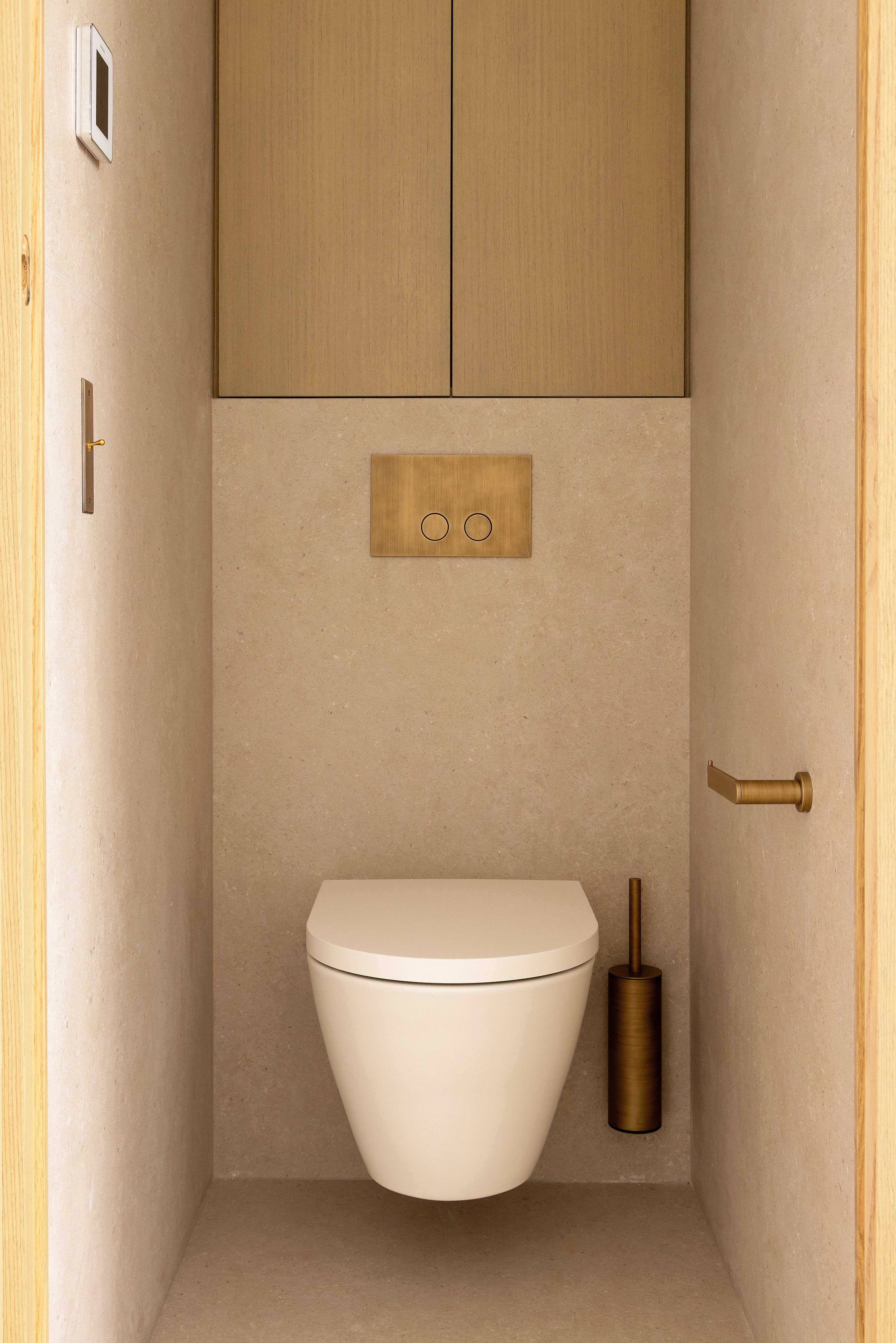 Toilettes suspendues avec plaque de chasse doree, revetement pierre beige et rangement en bois clair
