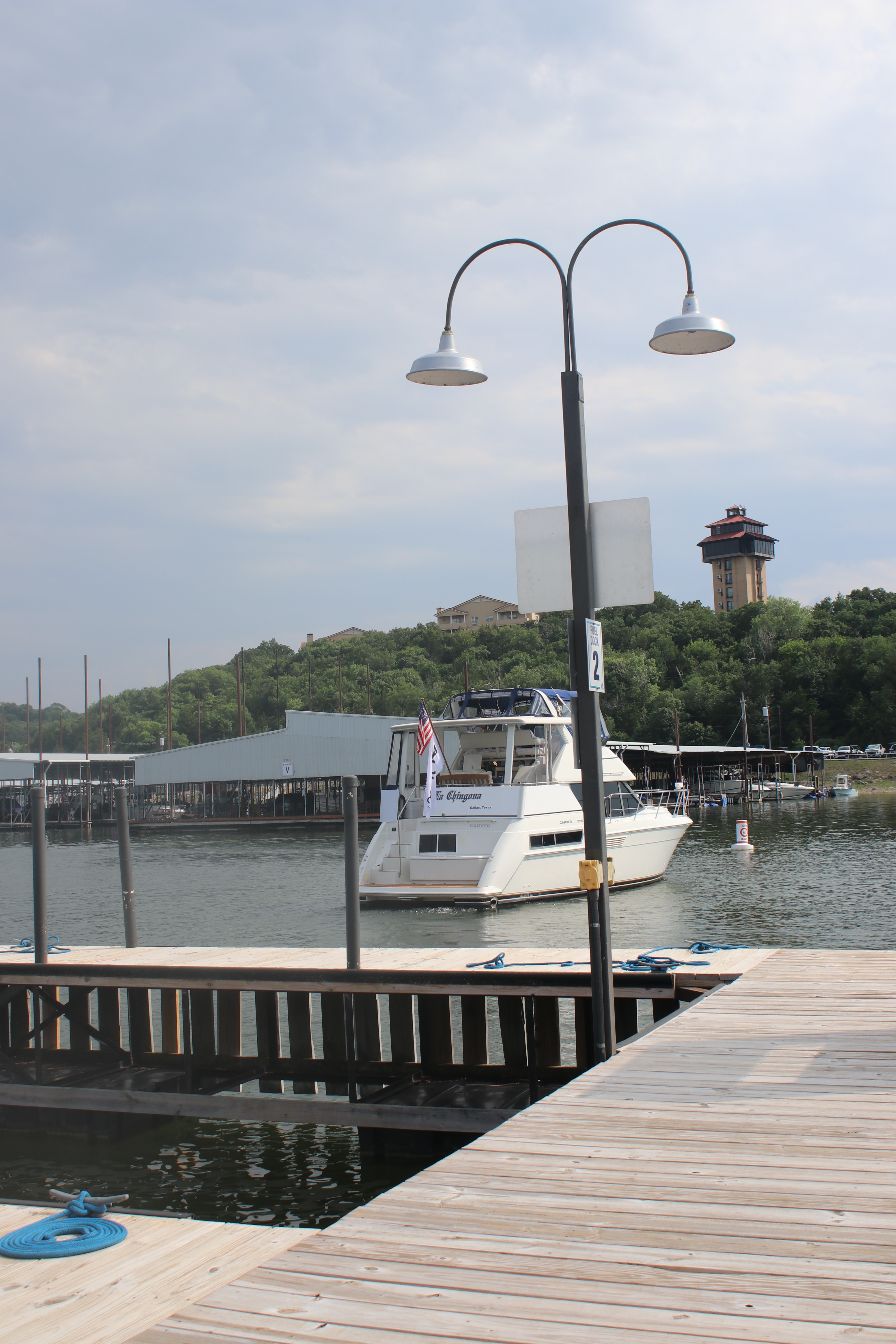 A white motor yacht is docked at a wooden marina, with a hilly landscape and buildings in the background under a partly cloudy sky.