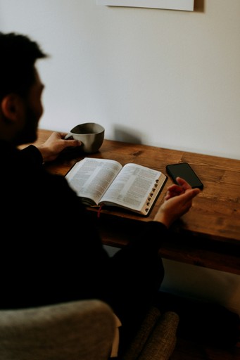 Personne assise à une table en bois, tenant une tasse de café dans une main et un téléphone portable dans l'autre, avec un livre ouvert devant elle. L'image est encadrée avec des coins arrondis.