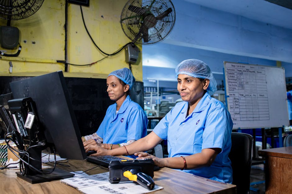 Female factory workers at desks using computers in an industrial setting, illustrating modern blue-collar employment.