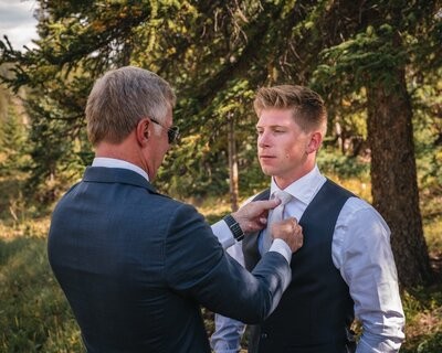 A man straightens Tucker's tie as the groom stands in his shirt and waistcoat.