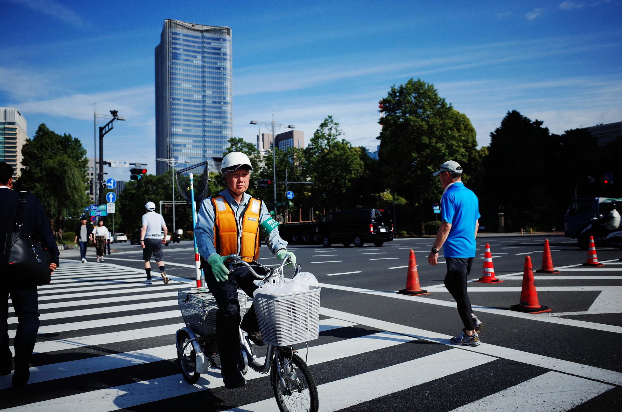 Photographie de rue prise sur un passage piéton d'un travailleur japonais sur son vélo et portant un gilet de sécurité orange vif et un casque blanc