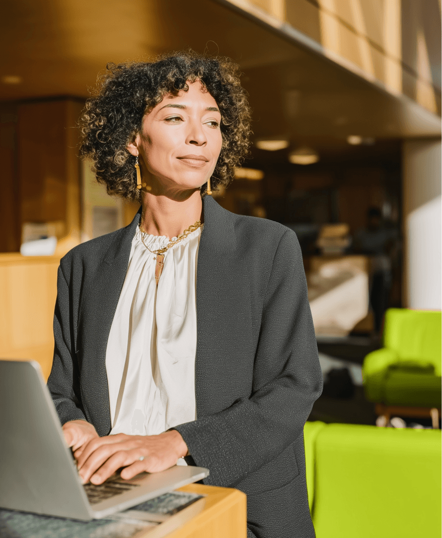 Foto de uma mulher com cabelos cacheados e blazer cinza, em pé na recepção de uma clínica bem iluminada com sofás verdes ao fundo. Ela está usando um notebook na Recepção da Clínica. A imagem simboliza a preparação antes de receber um paciente com o auxílio da Tivita.