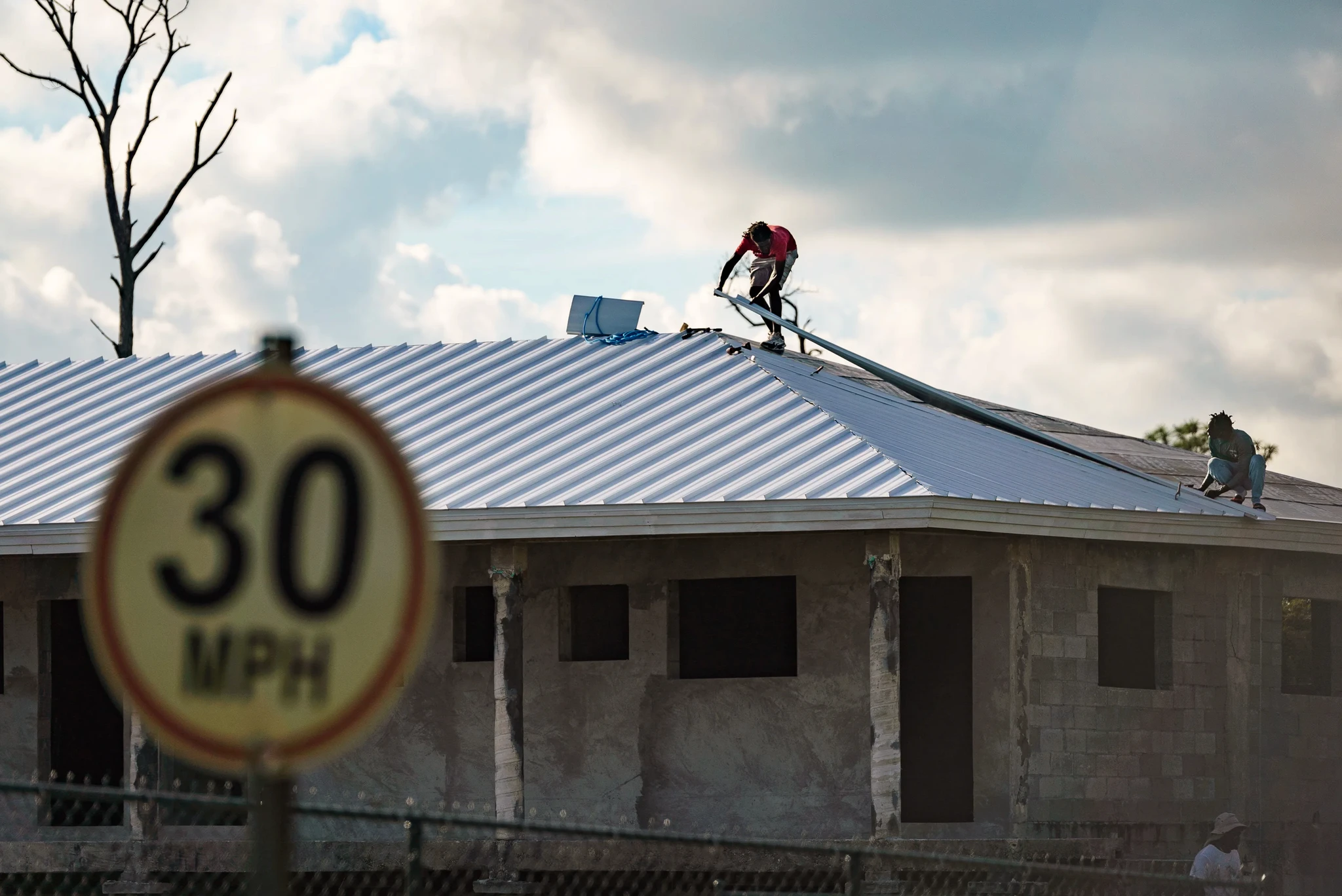 Two workers install metal roofing on a partially constructed house under a cloudy sky. A 30 MPH speed limit sign appears in the foreground.