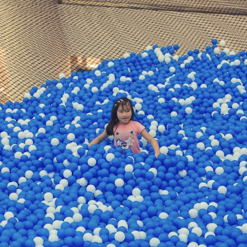 A young girl plays in a ball pit filled with blue and white balls, surrounded by a netted boundary.