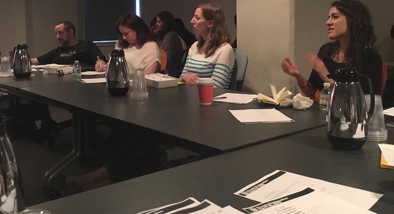 A photo of a group of people sitting around a table. On it there are cups, notepads and pens.