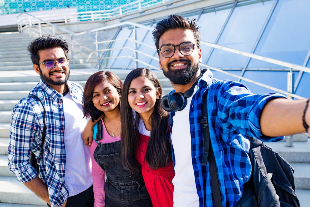 A group of four cheerful young friends, dressed casually, take a selfie together on a sunny day, standing in front of modern architectural steps, capturing their joyful camaraderie.