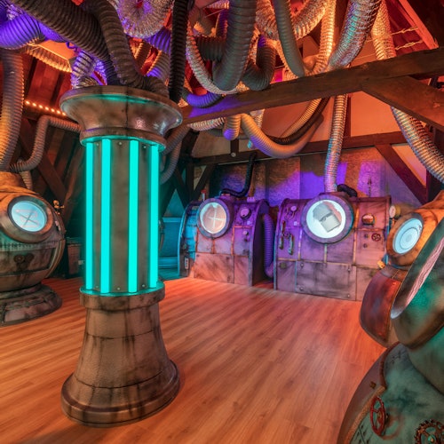 Futurisztikus szoba fém kapszulákkal, kör alakú ajtókkal, látható csövekkel és egy fénylő kék oszloppal. A fa padló kontrasztot alkot az ipari dekorációval.
