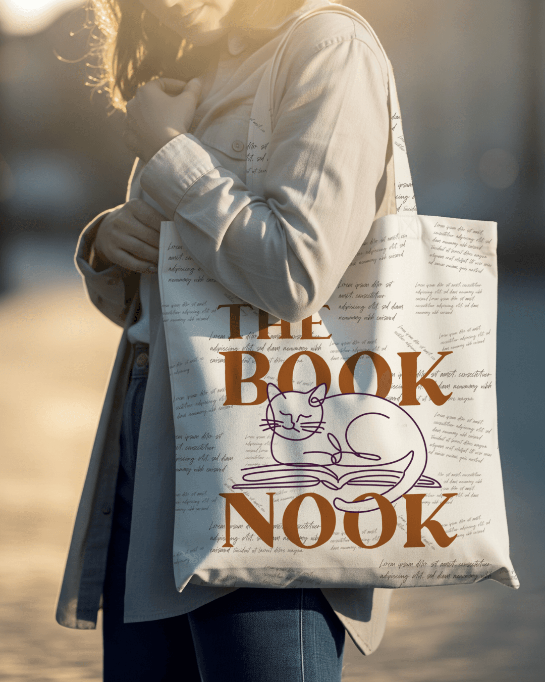 A canvas tote bag printed with THE BOOKNOOK in large amber serif letters and a purple line-art cat, with a pattern of handwritten script text covering the background, carried outdoors by a woman in a cardigan.