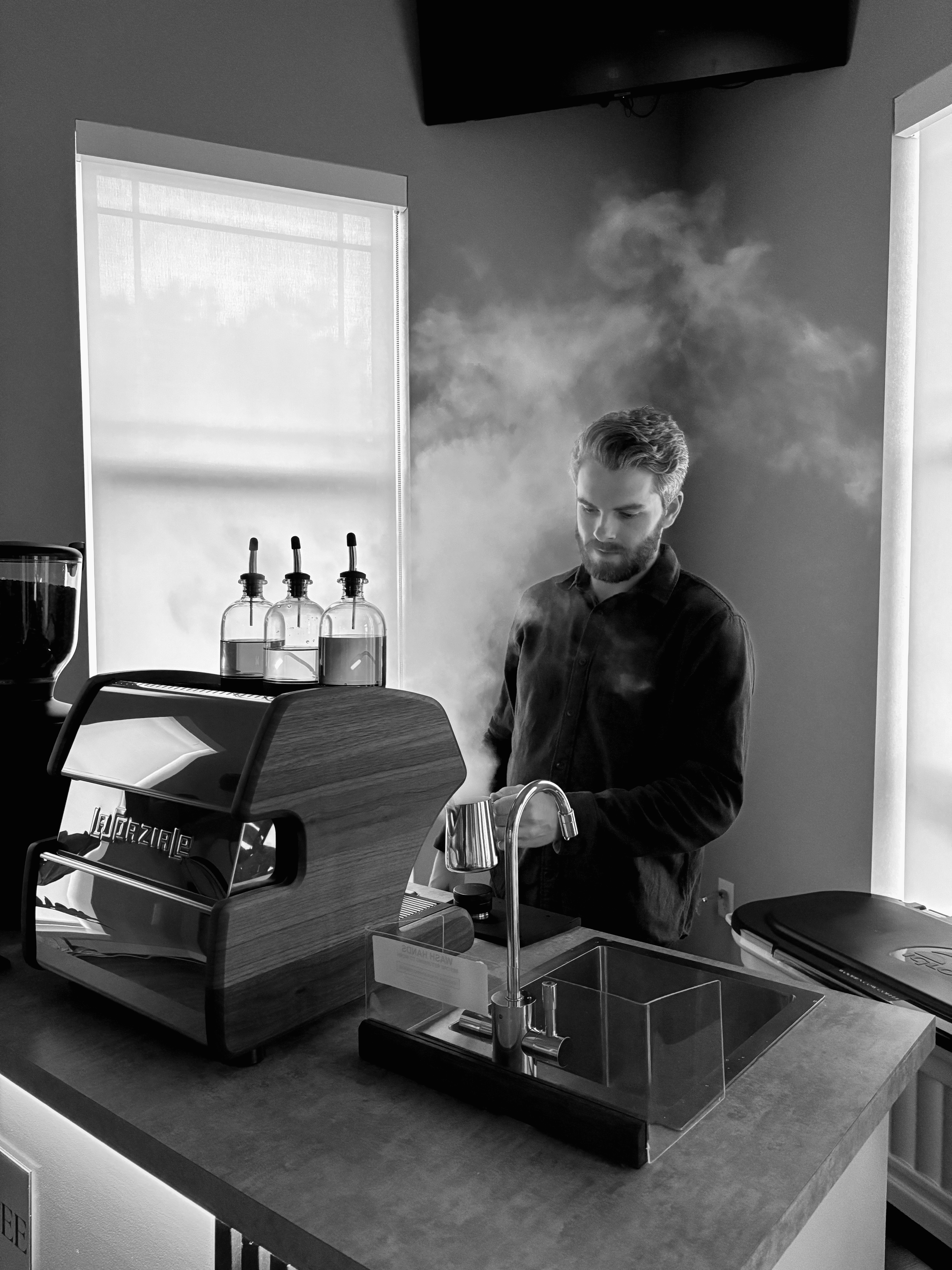 A man with a beard and neatly styled hair is steaming milk behind a modern espresso machine with a wood panel finish. Steam rises dramatically around him, filling the space. Three syrup bottles with amber liquid and dropper tops are placed on top of the machine. The setting appears to be indoors with diffused daylight coming through two tall windows with shades. The mood is calm and focused, capturing a moment of barista craftsmanship.