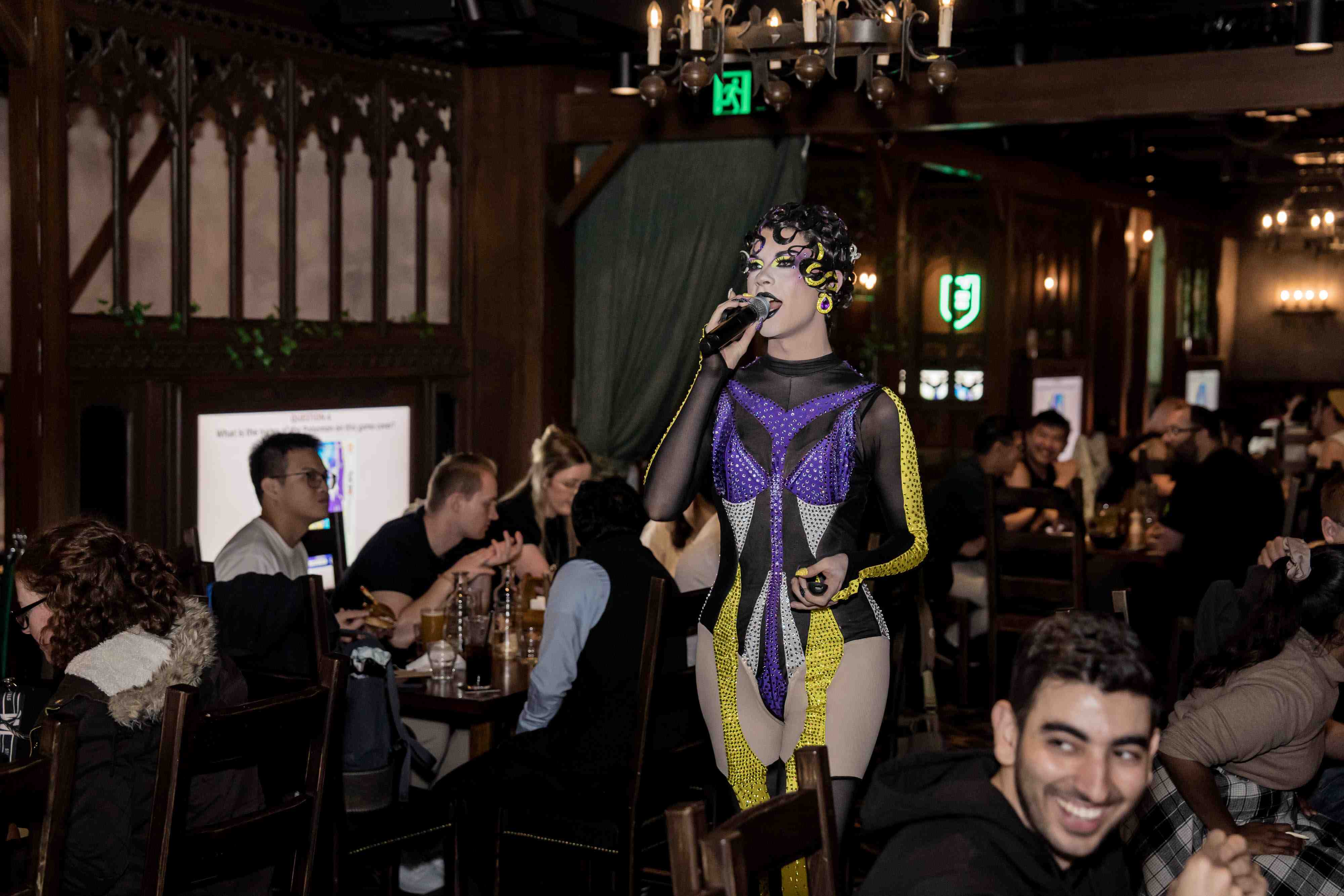 drag queen holding a microphone in a busy tavern while people smile