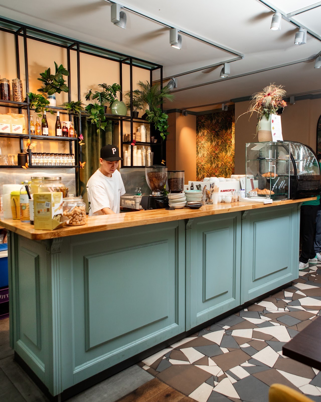 Ein Mann hält zwei farbenfrohe Getränke mit Garnierung in den Händen, er steht vor einer Theke mit einem Neon-Jederzeit-Schild und einem Kühlschrank.