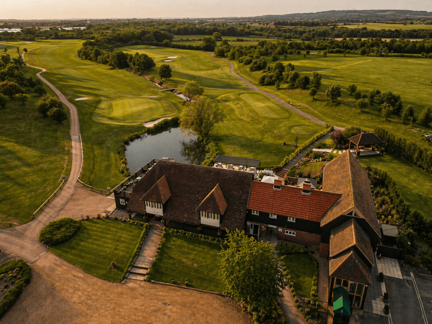 Aerial view of a golf course with a clubhouse featuring brown roofs, a pond, green grass, trees, and a path.