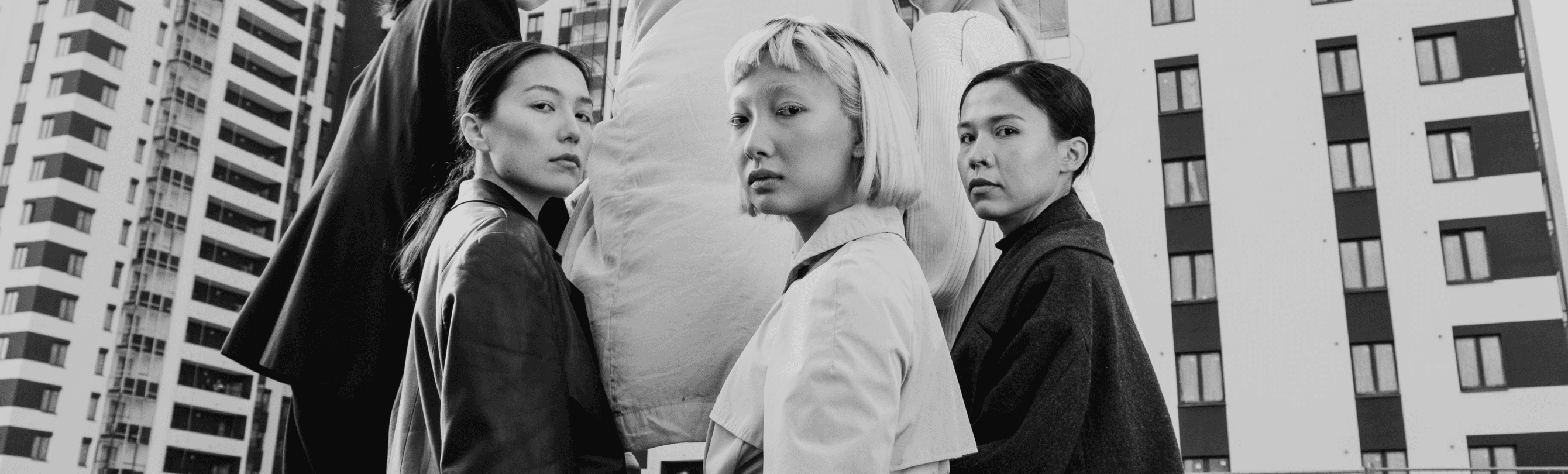 Three women with East Asian features stand closely together in front of a large, light object, posing outdoors against a backdrop of tall modern apartment buildings.