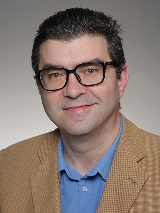 Portrait of a smiling man with glasses, a black shirt, and a gold chain. He has short dark hair.