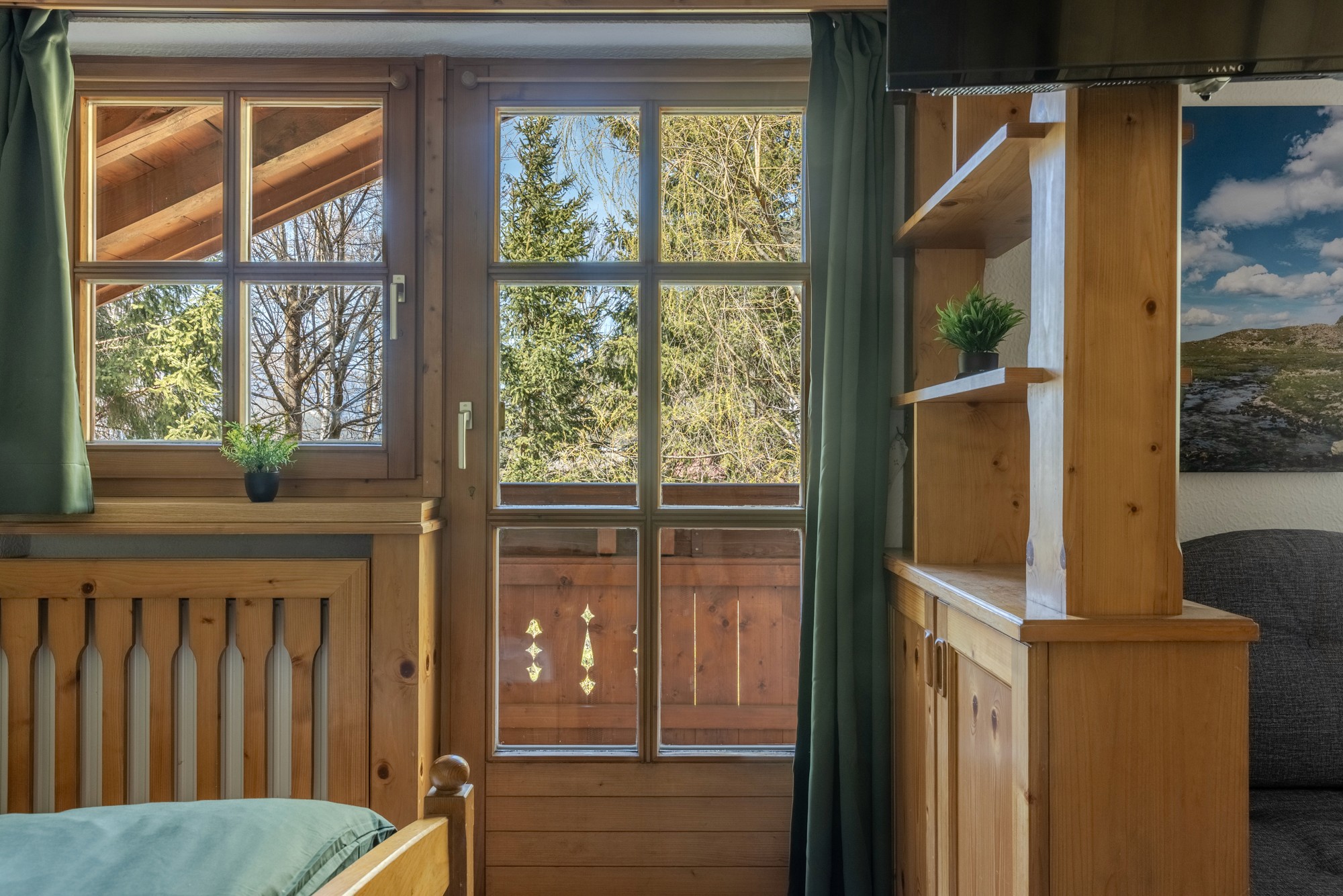 Ein gemütliches Interieur aus Holz mit einer Fenstertür, die auf einen Balkon führt und einen Blick auf üppiges Grün und Bäume bietet, wodurch eine ruhige, natürliche Atmosphäre entsteht.
