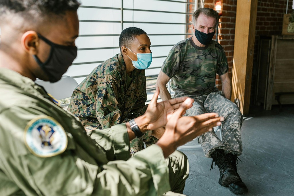 The image shows three military personnel wearing face masks engaged in a conversation. They are dressed in different military uniforms, indicating they might be from various branches of the armed forces. The setting appears to be indoors, with natural light streaming in through slatted blinds. The interaction seems informal and supportive, reflecting camaraderie and teamwork among the soldiers.