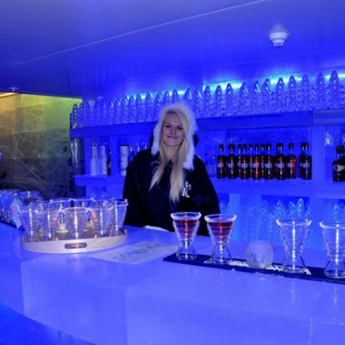 Человек с длинными светлыми волосами стоит за ледяной барной стойкой, окружённый бутылками, бокалами и синей подсветкой.