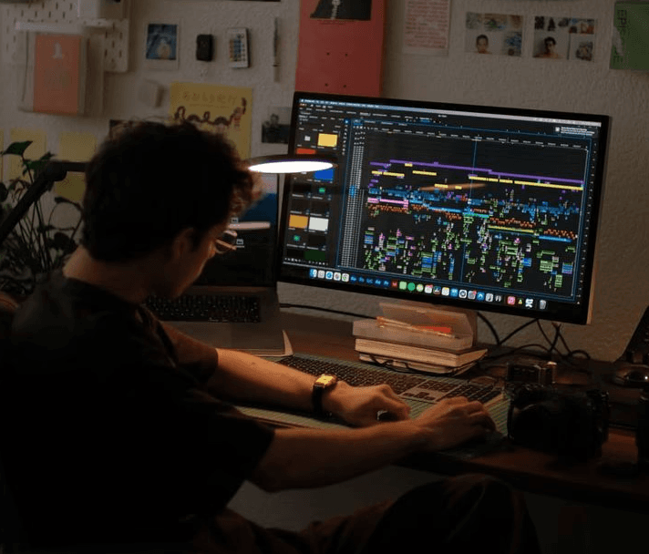 A smiling couple takes a selfie under a clear blue sky. The woman wears a red jacket, and the man, in a green top, holds a smartphone.