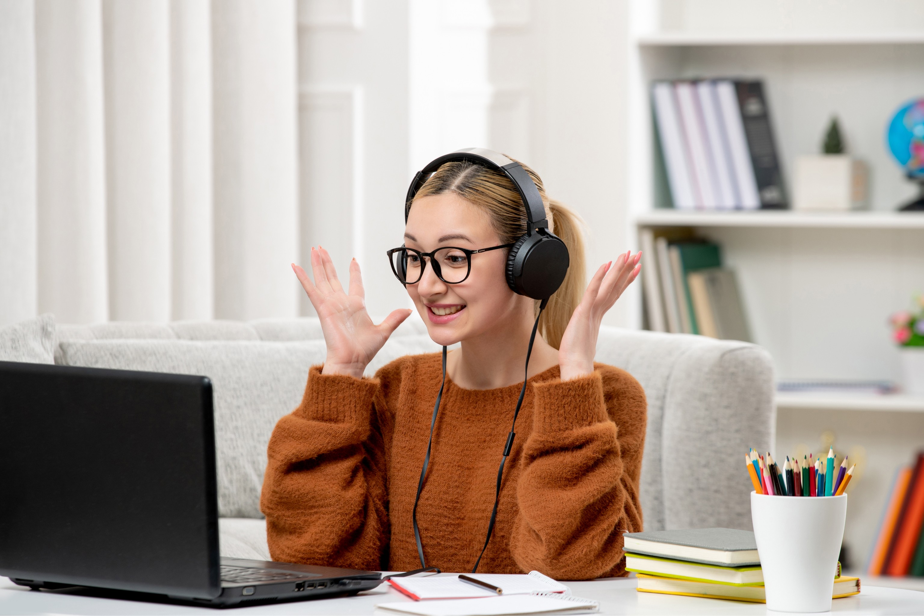 A person wearing headphones sits at a desk with a laptop, looking excited, surrounded by books, notebooks, and a cup of colorful pens, set in a cozy home office environment.