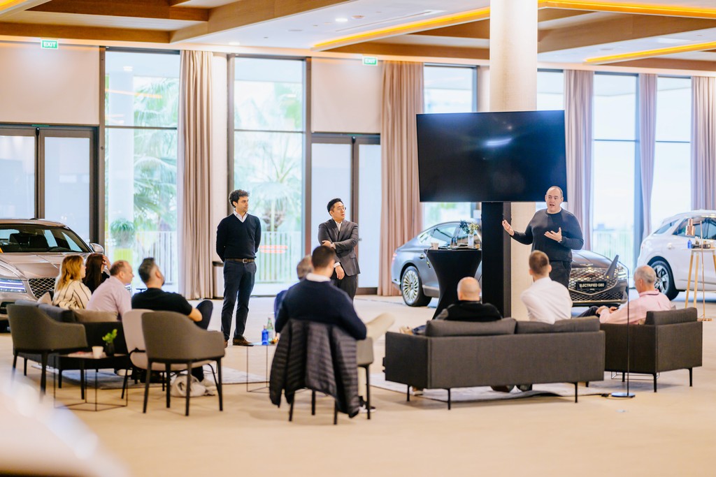 The speaker presents at a Genesis brand event in a modern showroom, surrounded by premium vehicles and an attentive audience.