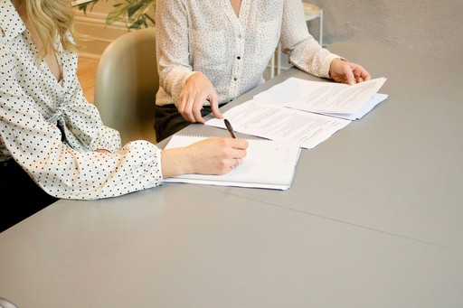 Two figures who are engaged in signing paperwork, in professional clothes in a professional setting