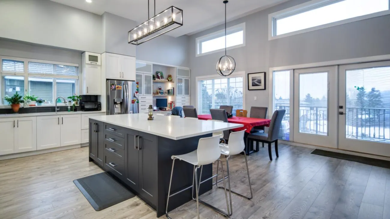 Gourmet custom kitchen in Burnaby home showcasing white perimeter cabinetry, oversized charcoal gray island with waterfall edge quartz countertops, pendant lighting, and open-plan dining area with abundant natural light