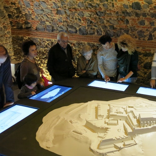 Isang grupo ng mga tao na may iba't ibang mga edad ay nagmamasid ng isang malaking modelo ng arkitektura at mga screen display sa isang silid na may mga dingding na bato.