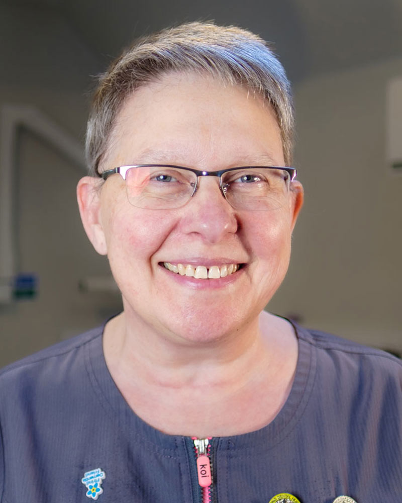 A portrait of Wendy, a Dental Nurse at Cricklade Dental Practice, smiling and wearing glasses and a dark grey scrub top with a pink zipper pull. The top features yellow embroidered text reading "WENDY" on the left and "Cricklade Dental Practice" on the right, along with several pinned badges. She is standing in a dental surgery with a dental light visible in the background.