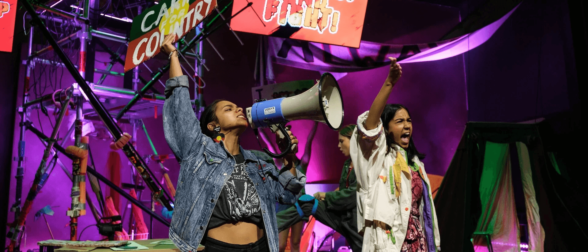 Two performers lead a protest chant under bright stage lights: one holding a “CARING FOR COUNTRY” sign and shouting into a megaphone, the other raising a fist. Behind them, a screen reads “STAND UP AND FIGHT!” with more performers and tents in the background.