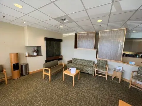 A bright, modern waiting room with beige chairs, wooden furniture, and a wall-mounted TV.