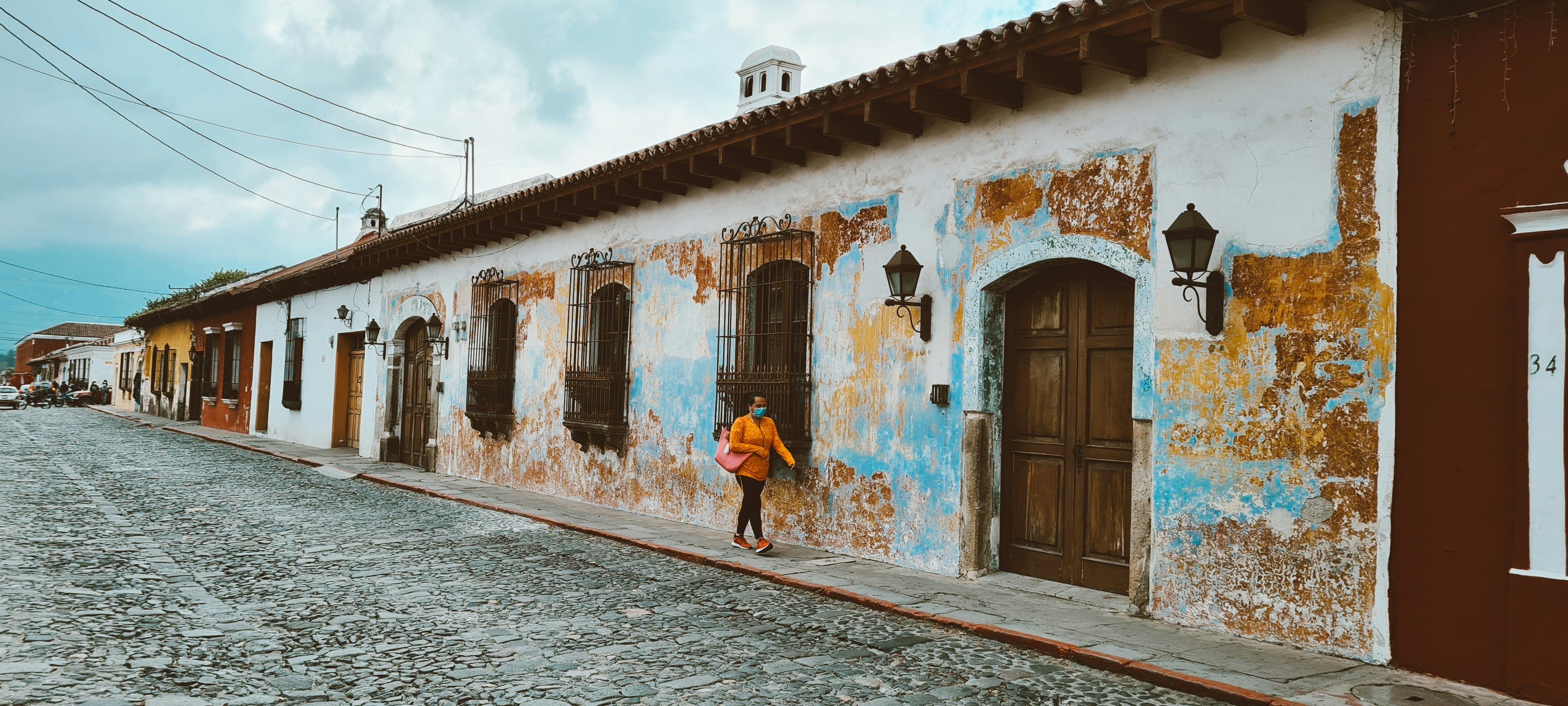 A person in a bright orange jacket walks along a cobblestone street with colorful, weathered stucco buildings, featuring wooden doors and iron-barred windows, in an old town setting.