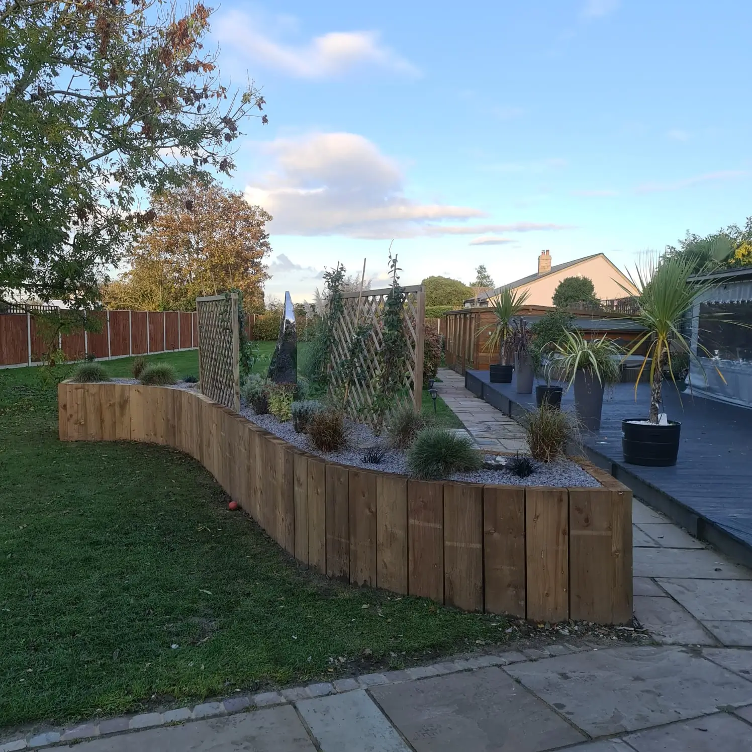A landscaped garden with a wooden fence, plants, and a pathway under a blue sky with scattered clouds.