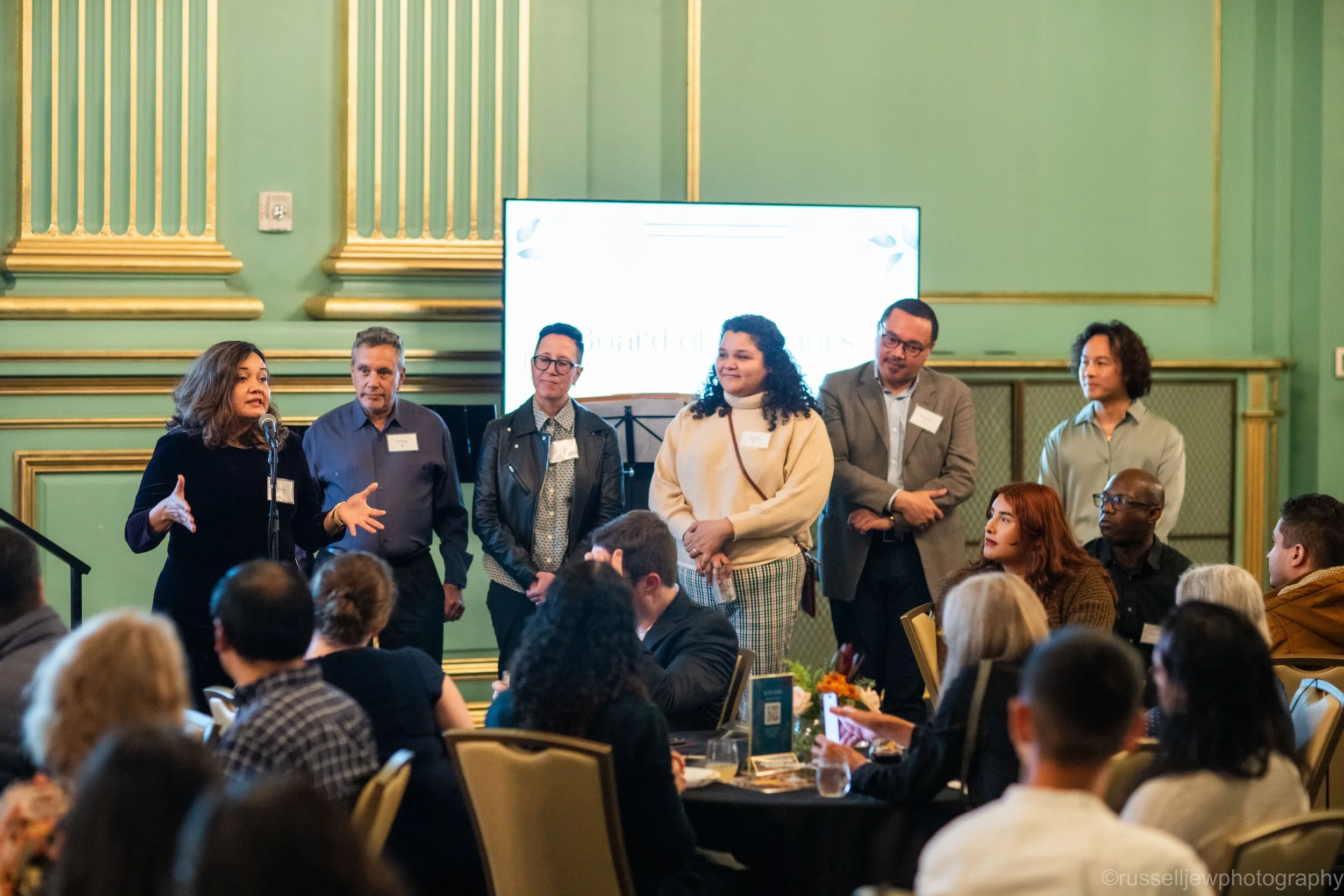 Um grupo de seis pessoas está na frente de um elegante salão de banquetes com paredes verdes, dirigindo-se a uma plateia sentada durante um evento formal.