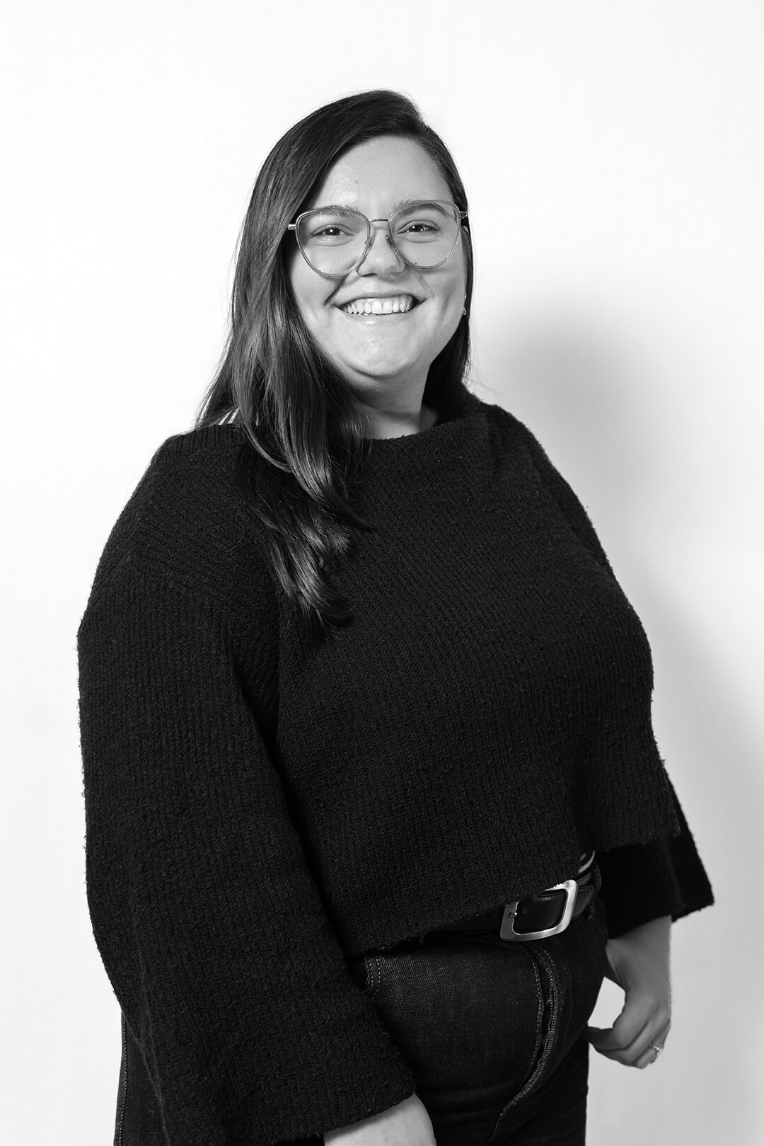 A smiling woman with glasses, wearing a dark sweater and jeans. She stands against a plain white background, exuding a friendly and confident demeanor.