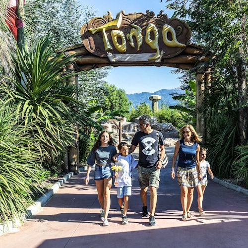 Viiden hengen perhe kävelee suuren puisen portin alla, jossa lukee "Tonga", vehreässä trooppisessa ympäristössä.