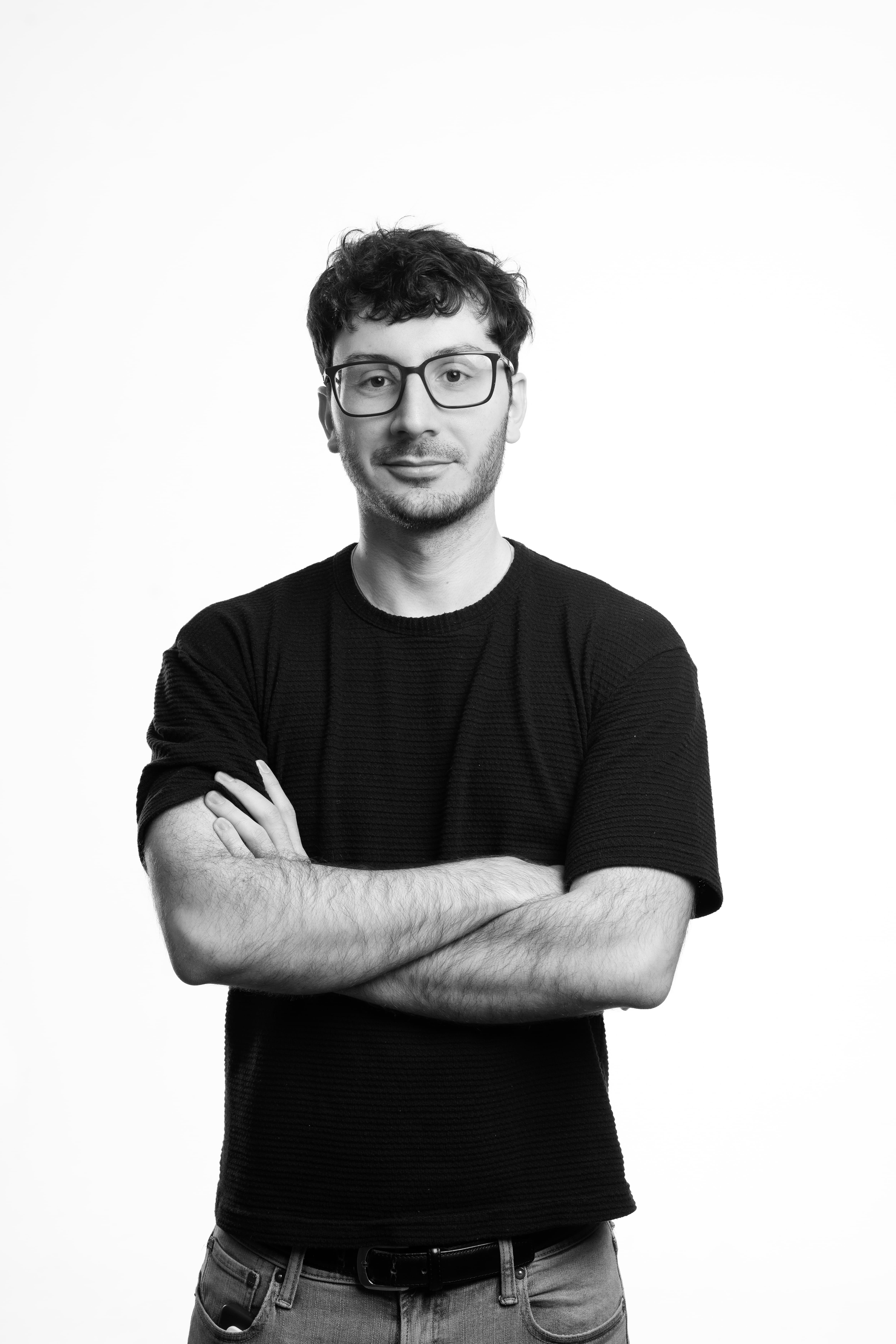 A person with glasses stands confidently with arms crossed, wearing a black T-shirt. The black-and-white image conveys a professional yet relaxed tone.