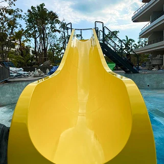 Một cầu trượt nước màu vàng sáng dẫn xuống khu vực bể bơi màu xanh lam, với một bộ cầu thang màu tối và một tòa nhà đang xây dựng ở phía sau dưới bầu trời một phần có mây.