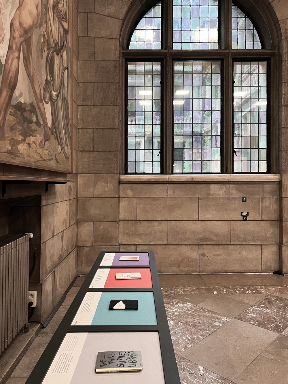 Een overzicht van de tentoonstelling in de KU Leuven bibliotheek. Op de voorgrond staat een tafel met een selectie boeken die geselecteerd zijn voor deze expo, met daarachter de indrukwekkende historische raampartijen van de bibliotheek.