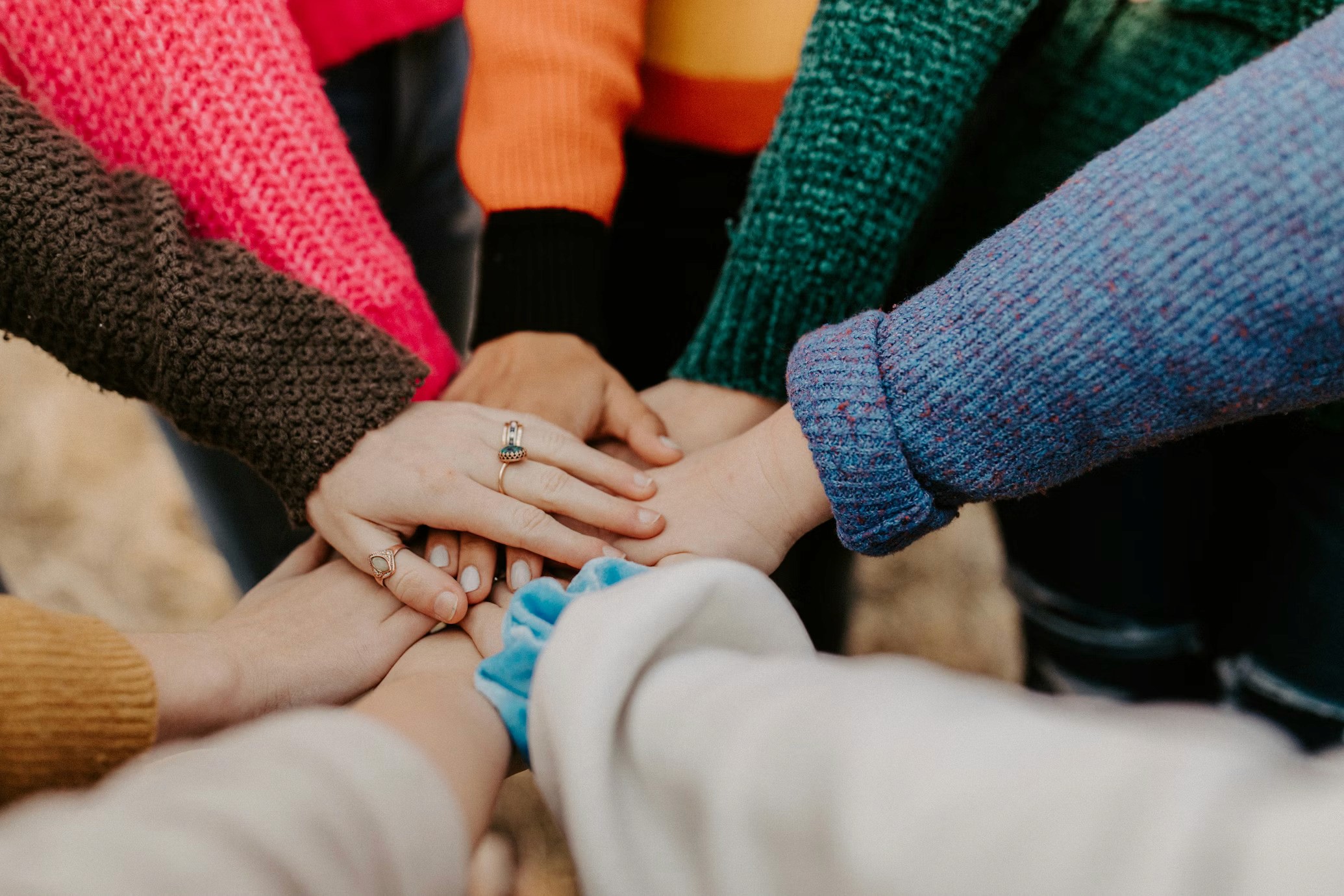 A close-up shot of several diverse hands stacked on top of one another in the centre of the frame, symbolising unity and collaboration. The people are wearing various colourful, textured sweaters in shades of pink, orange, green, and blue.