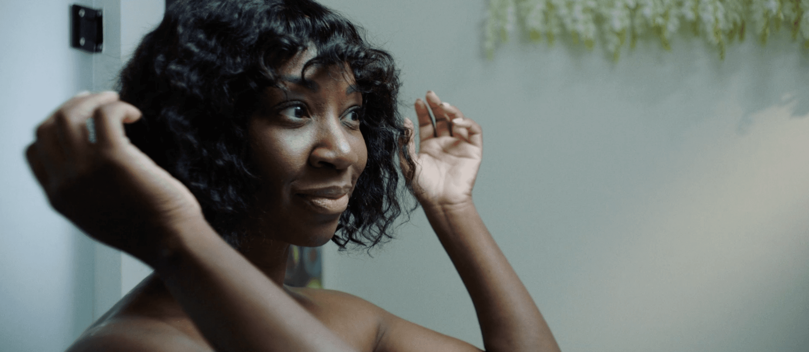 Natasha Ofili as Amara/Sade (a dark-skinned Black woman) adjusting their curly hair with a slight smile in a softly lit bathroom.