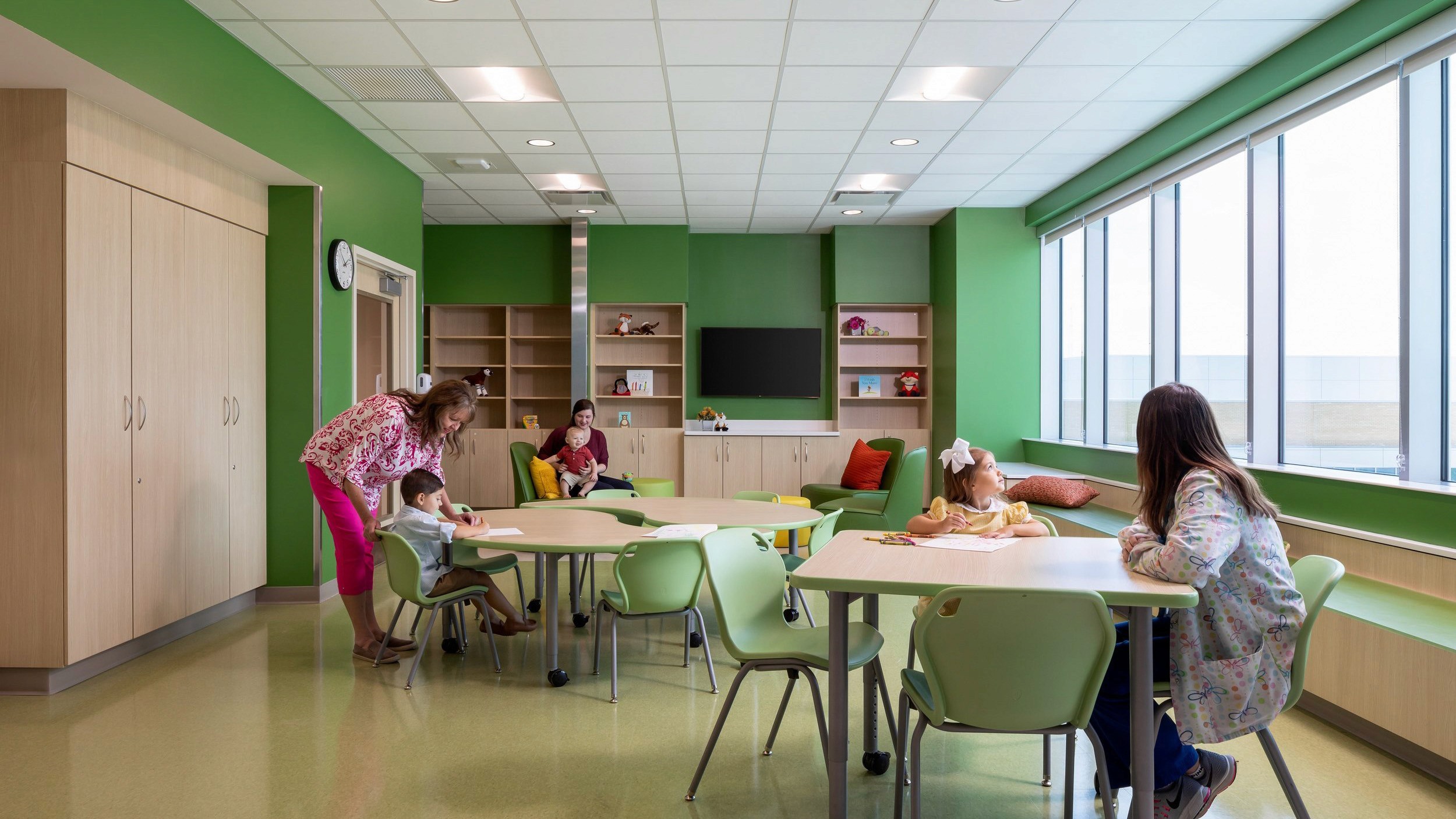 Children and adults in a bright room with green walls and light wood cabinets.
